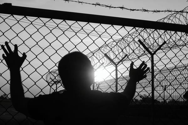 Migrant looking outside of prison fence: Original image licensed from Adobe Stock; grayscale applied by author